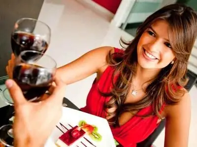 Woman in a red dress smiling and clinking wine glasses with another person at a restaurant table with dessert.