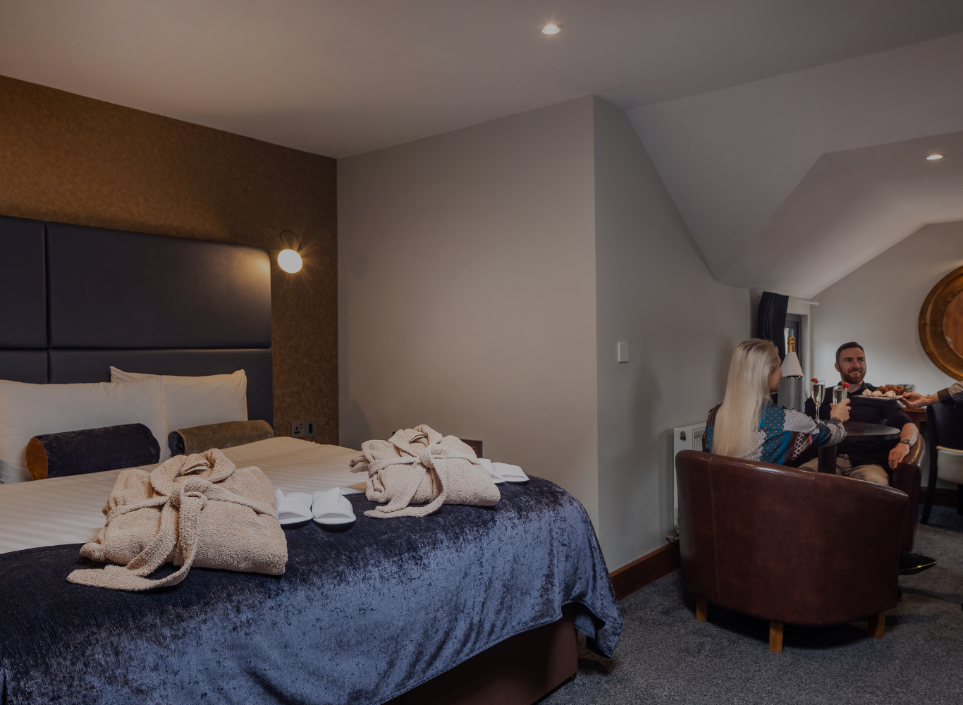 Elegant hotel room with king-sized bed, plush robes, and guests enjoying room service in a cosy seating area. Perfect for luxury stays.