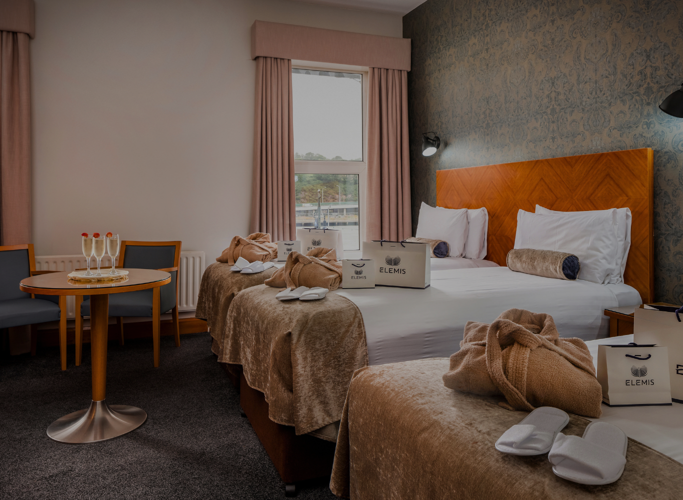 Elegant hotel room with two double beds, luxury Elemis amenities, cosy seating, and scenic window view, offering comfort.