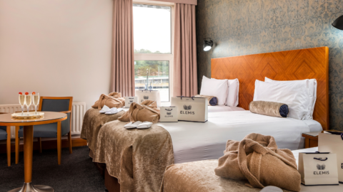 Elegant hotel room with twin beds, soft lighting, luxury Elemis toiletries, and a table with champagne near the window.