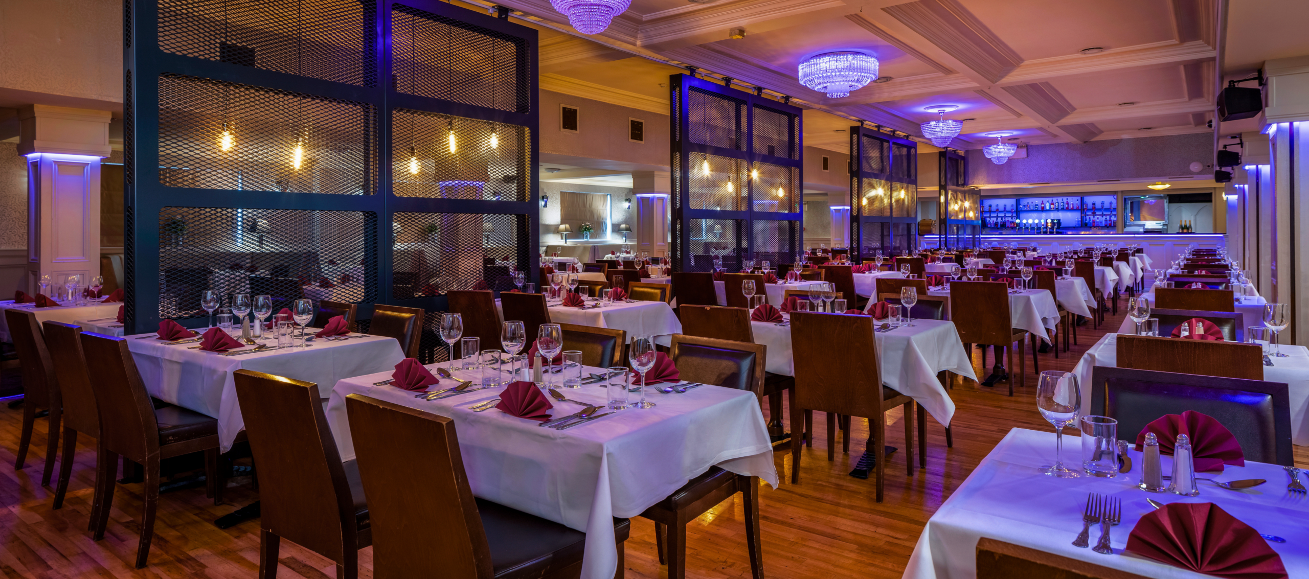 Elegant hotel dining room with ambient lighting, chic chandeliers, neatly set tables with burgundy napkins, and a stylish bar.