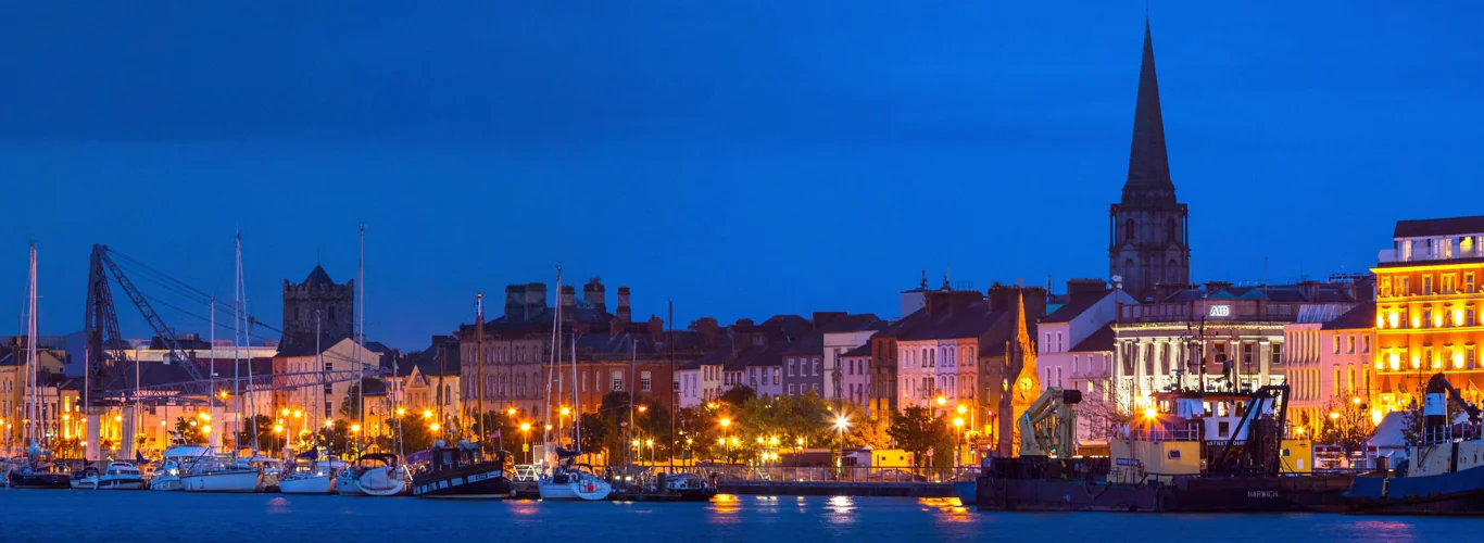 Vibrant city waterfront at twilight with boats, historic buildings, and glowing lights near the hotel for a scenic stay.