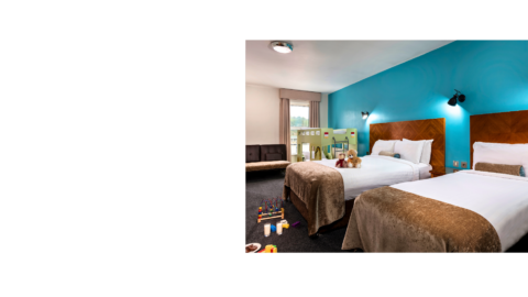 Modern hotel family room with two cosy double beds, soft toys, crib, playful decor, and natural light from large window.