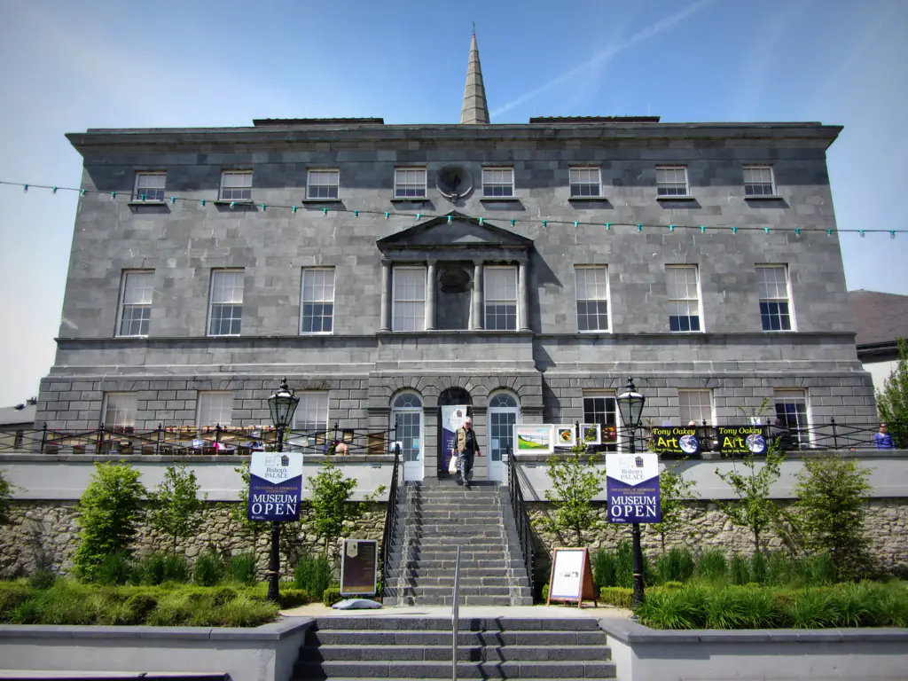 Bishops Palace Elegant stone hotel facade with outdoor seating, lush greenery, and inviting museum signs in sunny weather.