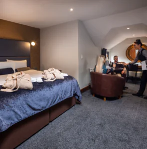 Elegant hotel room with plush bed, cosy robes, and guests enjoying room service at seating area under soft lighting.
